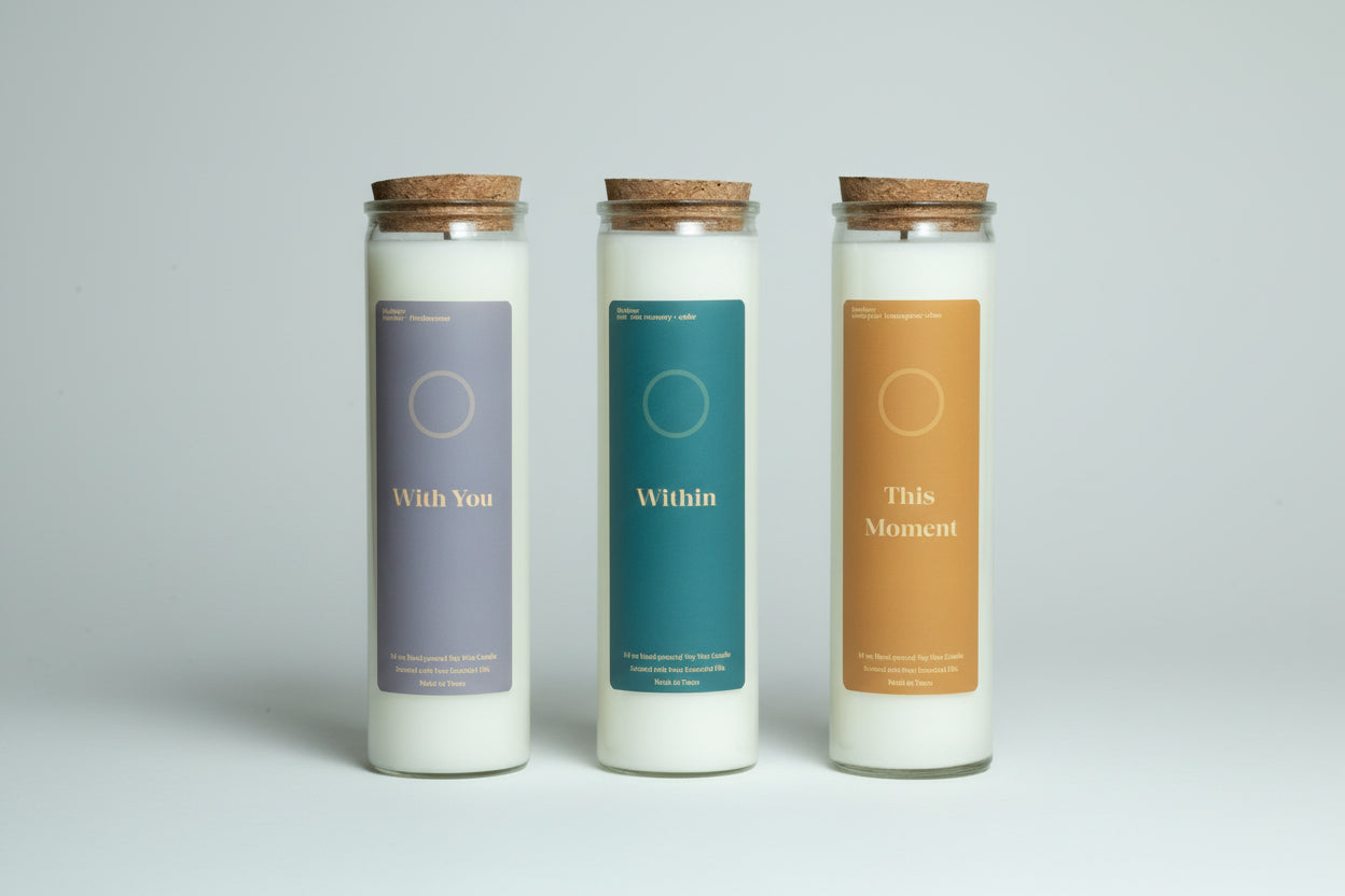 Three candles with labels 'With You', 'Within', and 'This Moment' on a surface with books and decor.