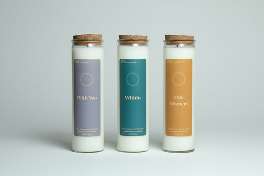 Three candles with labels 'With You', 'Within', and 'This Moment' on a surface with books and decor.